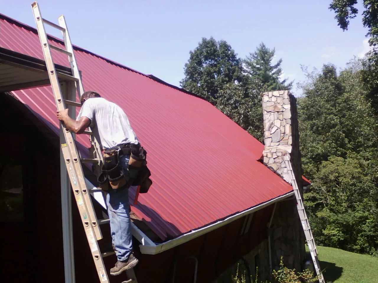 Roofer on a ladder beside a red metal roof and stone chimney — Soffit & Fascia Repair by Roofing Brothers in Seaside Heights